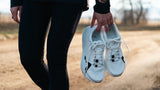 A person in athletic wear holds a pair of white running shoes outdoors.