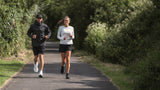 A man and woman running with running gear on a tree-lined country path.